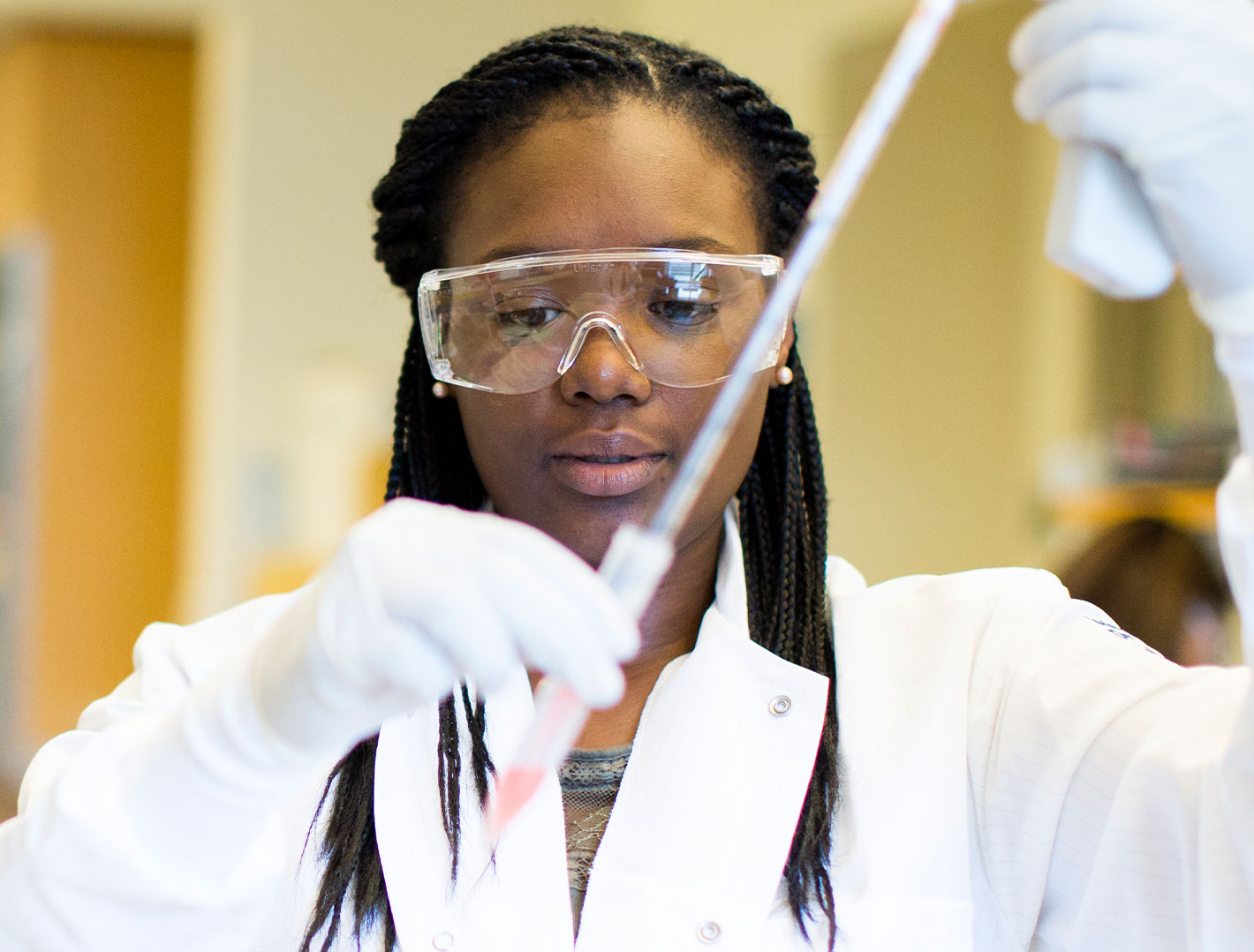 Student with a lab coat and goggles on holding a pipet in a lab setting.